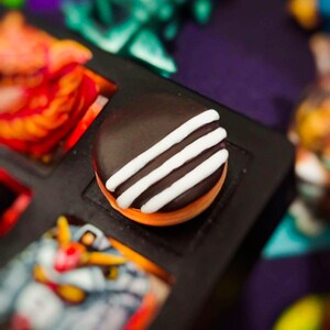 Op de afbeelding: Een rond, met donkerbruine chocolade bedekt koekje met drie parallelle witte glazuurstrepen aan de bovenkant. Het koekje heeft een zichtbare oranje basislaag en is geplaatst in een zwart, rechthoekig vakje van een dienblad.