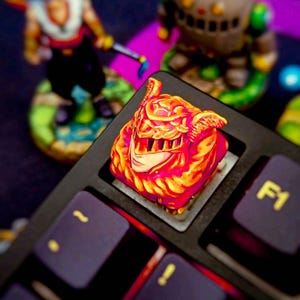 Pode incluir: Uma tecla de teclado personalizada com um design detalhado, laranja fogo e vermelho, do rosto de um personagem. A tecla está instalada em um teclado preto com teclas roxas. Outras figuras estão no fundo.