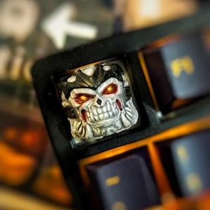 Può includere: Un tappo per tasti a forma di teschio bianco e nero con occhi rossi luminosi. Il tappo per tasti è su una tastiera nera con altri tasti visibili.