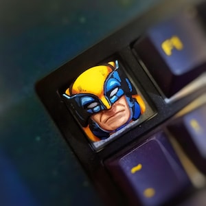 Pode incluir: Um tampão de tecla de teclado com uma ilustração amarela e azul do rosto do Wolverine. O tampão de tecla está em um teclado preto.