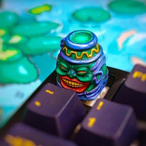 Può includere: Un tasto personalizzato per tastiera meccanica artigianale raffigurante un volto verde scolpito e dipinto a mano con una bocca larga e sorridente, labbra rosse e denti gialli, sormontato da una struttura decorativa blu e oro su un tasto viola scuro.