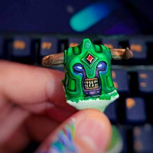以下が含まれることがあります： 手描きの緑色のアーティザンキーキャップ。角、赤い宝石、鋭い歯を持つ不気味な顔のヘルメットデザイン。キーキャップは暗い背景に対して保持され、その複雑なディテールと鮮やかな色を示しています。