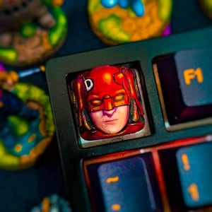 Pode incluir: Close-up de uma tecla de teclado personalizada com uma máscara de super-herói vermelha com a letra "D" na testa. A tecla faz parte de um teclado preto com teclas de função laranja. Outras figuras coloridas estão no fundo.