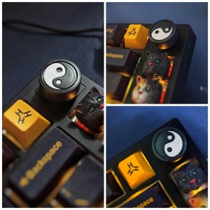 Pode incluir: Close-up de um teclado mecânico preto com teclas personalizadas. As teclas incluem um símbolo yin-yang, teclas em forma de gato e teclas amarelas com um símbolo. O teclado está sobre um fundo escuro.