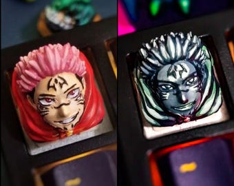 May include: Two custom keycaps featuring anime character designs. One keycap has a pink-haired character in a red cloak, the other has a gray-haired character. Both keycaps are detailed with facial features and markings.