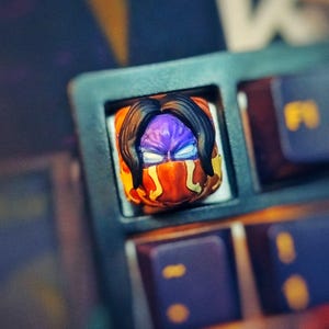 Pode incluir: Tecla de teclado artesanal personalizada com um rosto estilizado com cabelo preto, rosto roxo e detalhes em laranja e amarelo. A tecla está inserida em uma moldura de teclado preta.