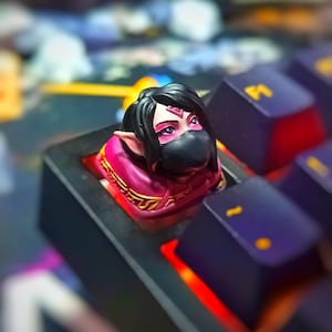 以下が含まれることがあります： 黒い髪、黒いマスク、ピンクのアクセントを持つキャラクターの、詳細な手描きのデザインが施されたカスタムアーティザンキーキャップ。 キーキャップは、紫色のキーと赤い照明を備えたメカニカルキーボードにセットされています。