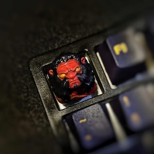 May include: A red and black 3D printed keycap featuring a lion's head with a fierce expression. The keycap is set against a black keyboard background.