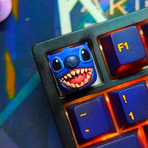 Pode incluir: Uma tecla azul personalizada com o personagem Stitch, com um rosto detalhado e boca aberta. A tecla está instalada em um teclado preto com teclas azuis e amarelas. A tecla F1 é visível.