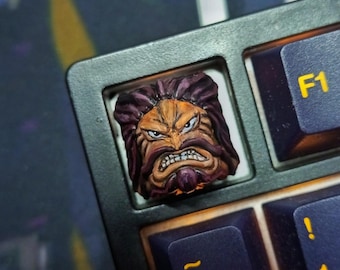 May include: Custom artisan keycap featuring a detailed, hand-painted face with a fierce expression. The keycap is brown, orange, and purple, set on a black keyboard with yellow lettering. The keycap is a unique addition to any mechanical keyboard.