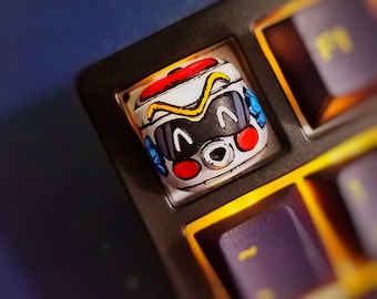 May include: A custom keycap featuring a cartoon character design. The keycap is white with a black face, red circles, and blue accents. The design is set on a black keyboard with purple keys.