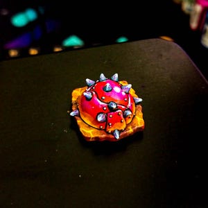Pode incluir: Uma tecla artesanal vibrante, pintada à mão, com um design vermelho e pontiagudo com detalhes prateados. A tecla assenta numa base castanha texturizada. A paleta de cores inclui tons de vermelho, rosa, laranja e prata, criando uma estética única e detalhada.