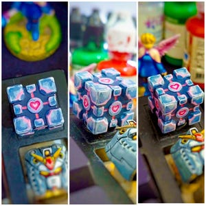 May include: A blue and pink cube-shaped figurine with heart designs. The cube has a dark blue base color with light blue and pink accents. The hearts are pink. The figurine is a collectible item.