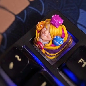 Op de afbeelding: Een aangepaste toetsenbordtoets met een handgeschilderd personage met geel haar, een paarse mantel en een kroon. Het personage wordt omringd door kleine, kleurrijke wezens.