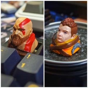 Puede incluir: Dos teclas personalizadas pintadas a mano con las caras de personajes populares de videojuegos. La primera tecla es un Kratos marrón y rojo de God of War, y la segunda tecla es una Aloy marrón y naranja de Horizon Zero Dawn.