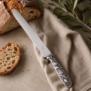Könnte beinhalten: Ein Brotmesser mit gezahnter Klinge und dekorativem Griff mit floralen und Blattmotiven. Das Messer aus Edelstahl liegt auf einem beigen Leinentuch neben einem Brotlaib und einer Scheibe.