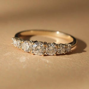 May include: A gold band ring featuring a series of six round-cut diamonds. The diamonds are set closely together, creating a continuous sparkle. The ring is set against a warm, neutral background, highlighting its elegant design.
