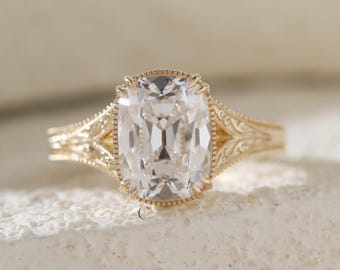 May include: A gold ring featuring a large, clear, cushion-cut center stone held by prongs. The band is crafted from yellow gold with intricate, engraved scrollwork patterns along the sides, leading up to the central setting.