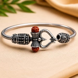May include: A silver-toned metal bangle bracelet featuring a flexible snake chain band. The centrepiece includes a silver trident symbol, a cylindrical bead with intricate engravings, and two small, round, textured reddish-brown beads.