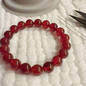 May include: A circular bracelet composed of numerous polished, deep red spherical beads strung together. The beads exhibit a translucent quality with subtle internal variations in colour and texture, arranged in a continuous loop on a white surface.