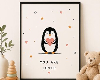 May include: A framed wall art print featuring a black and white penguin holding a pink heart. The background is off-white with scattered orange stars, black dots, and small pink hearts. The text reads YOU ARE LOVED in black capital letters.