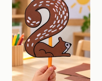 May include: A brown paper cutout shaped like the number two, designed to resemble a squirrel with a long, curved tail featuring white painted dashes. The cutout is attached to a solid orange stick, creating a handheld prop for educational use.
