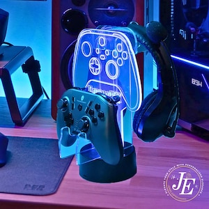 May include: A gaming accessory stand with a blue LED glow. The stand holds a black controller, a headset, and a clear acrylic controller-shaped holder. The stand has the text "JE" and "ELEVATE YOUR STYLE WITH PERSONALIZED ELEGANCE."