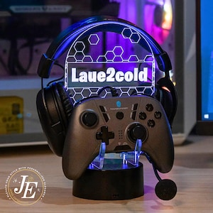 May include: A gaming headset and controller rest with a glowing blue and white hexagonal design. The stand features the text "Laue2cold" and a gray controller with the word "Thunderbot" on it. The base is black.