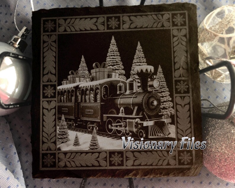 May include: A black square slate coaster with a detailed engraving of a vintage Christmas train. The train is surrounded by snow-covered pine trees and snowflakes. The text "Visionary Files" is at the bottom.