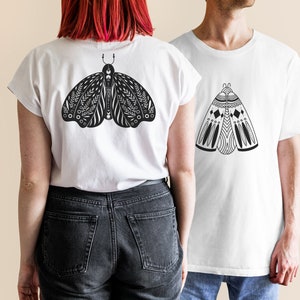 May include: Two white t-shirts with black and white moth designs. The moth on the left t-shirt has a more intricate design with floral elements. The moth on the right t-shirt has a more geometric design.
