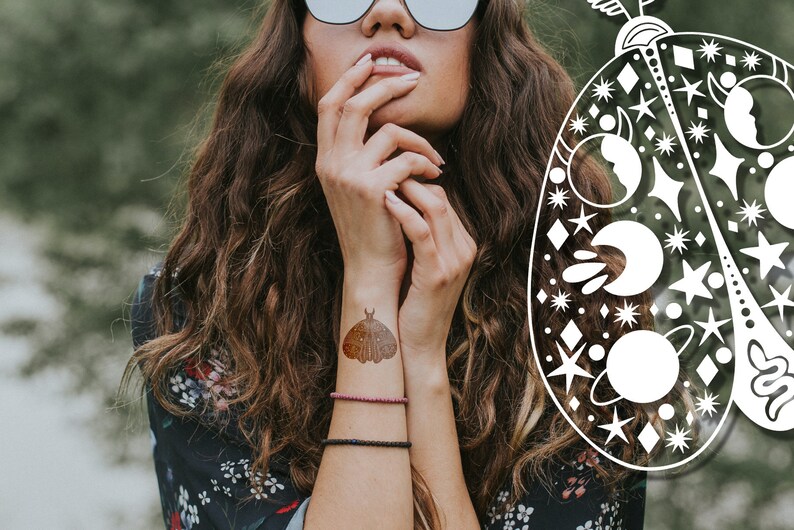 May include: A woman with long brown hair is wearing a floral patterned shirt and has a temporary tattoo of a moth on her wrist. The tattoo is a brown and gold color and has a celestial design.