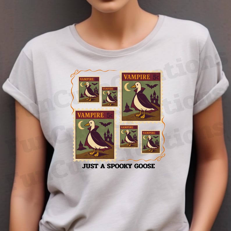 Op de afbeelding: Lichtgrijs t-shirt met een ontwerp van postzegels met een vampiergans-illustratie. De postzegels zijn in verschillende formaten en bevatten de tekst "VAMPIRE" en "JUST A SPOOKY GOOSE". De gans draagt een cape en staat voor een spookachtig kasteel.
