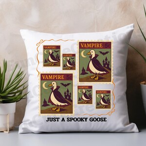 Op de afbeelding: Wit vierkant kussen met een print van een gans verkleed als vampier op postzegels. De postzegels tonen een gans met een cape, een halve maan, vleermuizen en een kasteel. Tekst luidt "VAMPIRE 5c" en "JUST A SPOOKY GOOSE."