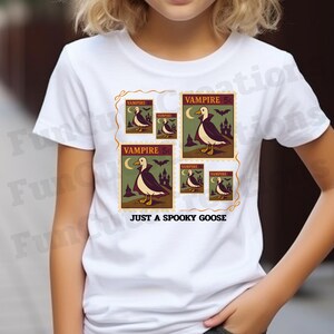 Op de afbeelding: Wit T-shirt met een grafisch ontwerp van postzegels in vintage stijl. De postzegels tonen een vampiergans met een cape, een halve maan en de teksten "VAMPIRE" en "JUST A SPOOKY GOOSE". Het ontwerp heeft een retro esthetiek.