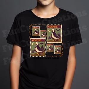 Op de afbeelding: Zwart T-shirt met een ontwerp van postzegels met een illustratie van een vampiergans. De postzegels hebben een waarde van 5 cent en bevatten de tekst "VAMPIRE" en "JUST A SPOOKY GOOSE". Het ontwerp bevat vleermuizen, een maan en een kasteel.