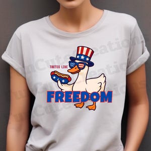 May include: A light gray t-shirt featuring a cartoon duck wearing sunglasses and a patriotic hat, holding a hot dog. The text "TASTES LIKE" and "FREEDOM" are also displayed in red and blue.