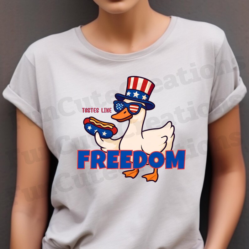 May include: A light gray t-shirt featuring a cartoon duck wearing sunglasses and a patriotic hat, holding a hot dog. The text "TASTES LIKE" and "FREEDOM" are also displayed in red and blue.