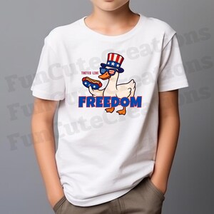May include: White t-shirt featuring a cartoon duck wearing sunglasses and a patriotic hat, holding a hot dog. The word "FREEDOM" is displayed in blue block letters below the duck. The text "TASTES LINE" is above the hot dog.