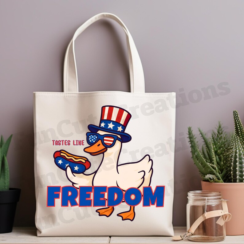 May include: A cream-colored tote bag featuring a cartoon goose wearing sunglasses and a patriotic hat, holding a hot dog. The text "TASTES LIKE FREEDOM" is displayed in red and blue.