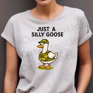 May include: A pale grey t-shirt with the words "JUST A SILLY GOOSE" above a cartoon goose. The goose is wearing a camouflage hat and vest, with a white body, orange beak and feet, standing on green grass.