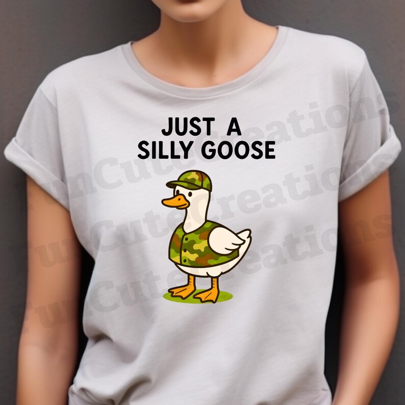 May include: A pale grey t-shirt with the words "JUST A SILLY GOOSE" above a cartoon goose. The goose is wearing a camouflage hat and vest, with a white body, orange beak and feet, standing on green grass.