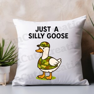 May include: White square cushion featuring a cartoon goose in a camouflage outfit, with the text "JUST A SILLY GOOSE". The goose has a yellow beak and feet, and is standing on green grass.