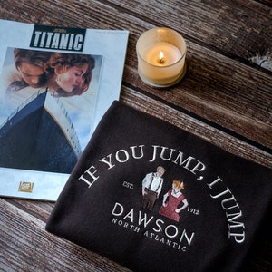 Puede incluir: Una camiseta negra con el texto "If you jump, I jump" y las palabras "Dawson North Atlantic" debajo. La camiseta también presenta una pequeña ilustración de dos personas de pie una al lado de la otra. Una revista con la imagen de portada de la película Titanic está en el fondo.
