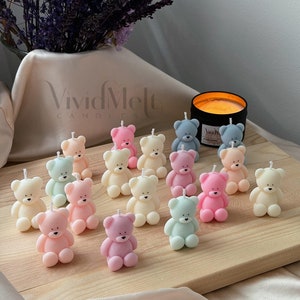 Op de afbeelding: Een verzameling van 15 pastelkleurige teddybeerkaarsen op een houten oppervlak. De kaarsen zijn in een halve cirkel gerangschikt met een zwarte kaarsenpot op de achtergrond. De kaarsen zijn allemaal in verschillende kleuren, waaronder roze, blauw, groen, geel en oranje.