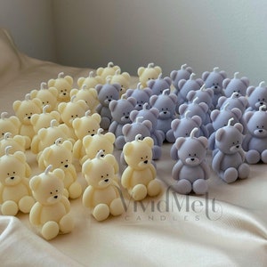 Pode incluir: Uma coleção de pequenas velas em forma de urso de pelúcia em duas cores: amarelo claro e roxo claro. As velas estão dispostas em filas sobre uma superfície bege claro. O texto "VividMelt CANDLES" é visível no canto inferior direito da imagem.