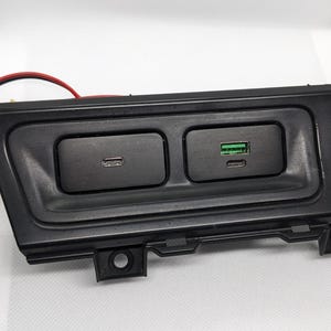 Puede incluir: Un panel de plástico negro con dos puertos de carga. El puerto izquierdo es un puerto USB-C y el puerto derecho es un puerto USB-A.