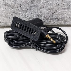 Puede incluir: Un micrófono negro para coche con una placa frontal rectangular con ranuras verticales. El micrófono está conectado a un cable negro y a un conector de audio de 3,5 mm. El cable está enrollado.