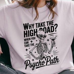 May include: A pale pink t-shirt with the slogan "Why take the high road? When you can take the Psycho Path." The design includes a skeleton with a cocktail. The person in the image is wearing sunglasses and a brown hat.