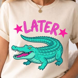 Puede incluir: Camiseta color crema con un diseño de un caimán turquesa y la palabra "LATER" en rosa. El caimán tiene la boca rosa y está rodeado de estrellas rosas. La camiseta tiene mangas cortas y un corte holgado, ideal para un look casual.