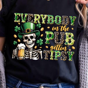 Op de afbeelding: Zwart T-shirt met een ontwerp voor St. Patrick's Day. De afbeelding toont een skelet met een groene leprechaun hoed, een biertje vasthoudend, en de tekst "EVERYBODY in the PUB gettin' TIPSY". Gouden munten en klavers zijn ook in het ontwerp opgenomen.